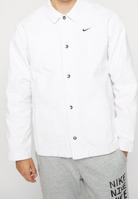 Person som bär en vit knäppbar Nike-jacka och grå sweatpants med svart "NIKE"-text på vänster lår, h änderna avslappnat vid sidorna.