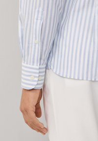 Lichtblauw en wit gestreept shirt met knoopmanchetten; gemaakt van stof met een gladde textuur; heeft een zak op de mouw.