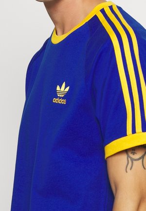 T-shirt bleu Adidas avec col et bords de manches jaunes, trois bandes jaunes sur les manches et logo Adidas jaune sur la poitrine.