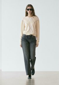 Maglione pesca chiaro con motivo a diamante, abbinato a jeans scuri a gamba dritta e scarpe nere. La modella indossa occhiali da sole neri oversize.