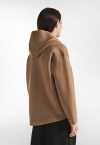 Bruine capuchontrui met een losse pasvorm, geribbelde manchetten en een zachte textuur. Gezien van achteren, die het hangende ontwerp van de hoodie toont.