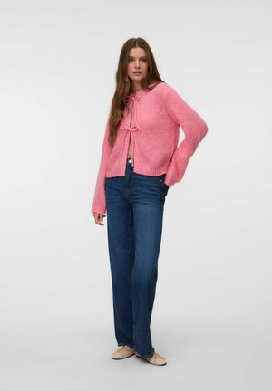 Femme portant un cardigan rose en maille noué à l'avant, un jean bleu taille haute et des chaussures plates beiges, debout devant un fond clair uni.