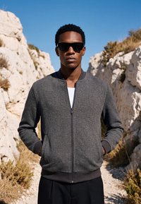 Un homme portant des lunettes de soleil foncées et une veste grise à fermeture éclair se tient les mains dans les poches entre des falaises rocheuses sous un ciel bleu clair.