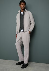 Gaiši pelēka bomber jaka virs tumši pelēka zip-up džempera, apvienota ar gaiši pelēkiem biksēm un melniem apaviem, uz neitrāla fona.