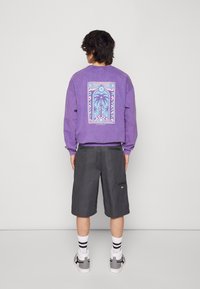 Felpa viola con stampa grafica di palme e accenti geometrici sul retro, abbinata a pantaloni corti grigi e calzini bianchi con strisce nere.