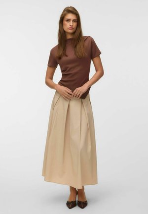 Jeune femme debout portant un haut marron à manches courtes, une jupe midi plissée beige et des ballerines pointues marron sur un fond uni.