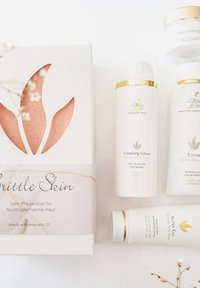 Fünf Hautpflegeprodukte in weißen Behältern mit goldenen Akzenten, die um eine weiße Box mit einem roségoldenen Design und Text angeordnet sind.