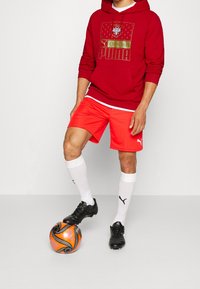 Felpa rossa con dettagli dorati, pantaloni corti rossi, calzini bianchi e scarpe da calcio nere, con un pallone da calcio arancione e nero su uno sfondo bianco.