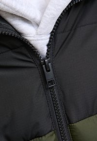 Czarna i zielona pikowana kurtka z widocznym zamkiem oraz szarym kapturem pod spodem, wykonana z miękkich w dotyku materiałów.