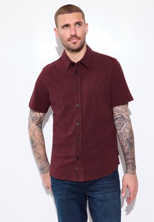 Man met korte baard en tatoeages op beide armen, draagt een bordeauxrood kortemouwen overhemd met knopen en donkerblauwe spijkerbroek, kijkt omhoog en naar rechts.