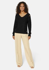 Schwarzer Strickpullover mit V-Ausschnitt, der über Aussparungen und gerippte Bündchen verfügt, kombiniert mit beigen weit geschnittenen Hosen und Sandalen mit offenen Zehen.