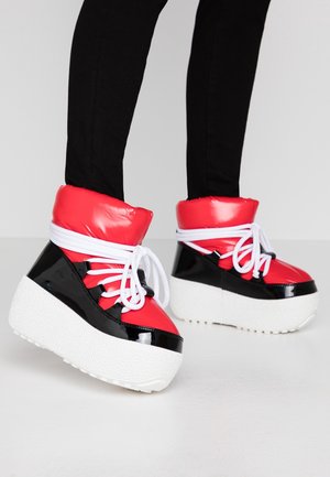 Jambes en pantalon noir portant des bottes d'hiver à plateforme rouges et noires avec des semelles épaisses blanches et des lacets blancs sur un fond uni.