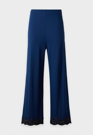 Femme aux longs cheveux foncés portant un top noir asymétrique et un pantalon large bleu marine taille haute avec des ourlets noirs ornés.