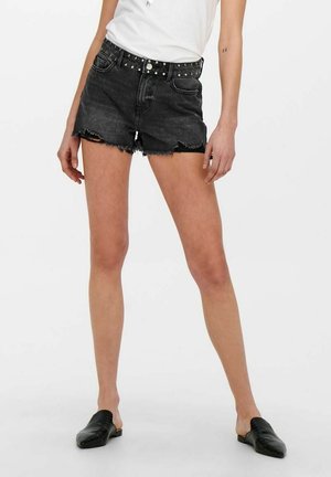 Person trägt schwarze, ausgefranste Jeansshorts mit Nietengürtel, weißes Oberteil und schwarze Slip-On-Schuhe und steht mit gespreizten Beinen.