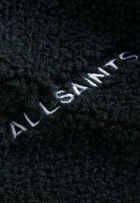 Juodas fliso audinys su tekstūru, kuriame matosi suktos skaidulos, akcentuotas balta siuvinėta prekės ženklo pavadinimu "ALLSAINTS".