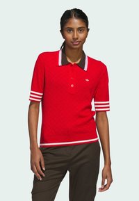 Polo rouge en tricot à manches courtes, avec un col marron et des accents rayés blancs. Présente un petit logo sur la poitrine.
