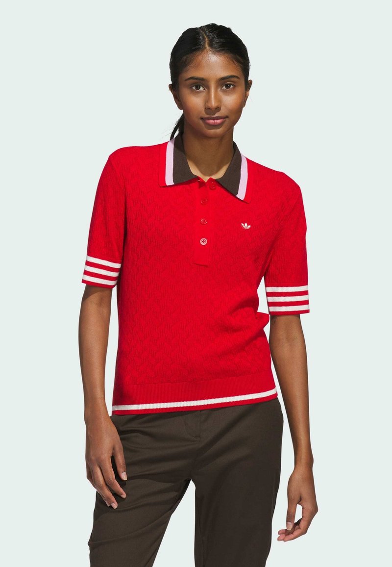 Polo rouge en tricot à manches courtes, avec un col marron et des accents rayés blancs. Présente un petit logo sur la poitrine.