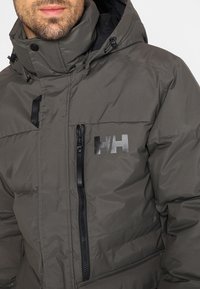 Giacca isolata grigia con colletto alto, chiusura a zip e accessori neri. Presenta il logo sulla tasca del petto e dettagli a coste.