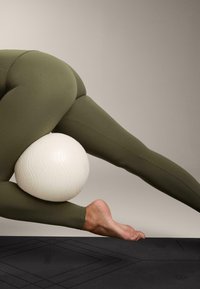 Olíva színű leggingset viselő személy, aki egy fehér, texturált jógagömböt egy fekete jógaszőnyegen a behajlított térd és az alkaron egyensúlyoz.