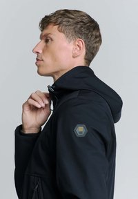 Zwarte hoodie met een gematteerde textuurpatch op de linker mouw. De stof lijkt mat, met een ritssluiting aan de voorkant en een verstelbare capuchon.