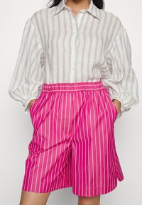 Bílé pruhované košile s knoflíky a volnými rukávy; v kombinaci s růžovými šortkami s vertikálními bílými pruhy a elastickým pasem.