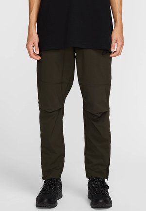 Pantalones cargo verdes de material ligero, con un corte relajado, paneles en las rodillas y detalles de cremallera en los tobillos, combinados con zapatos negros.