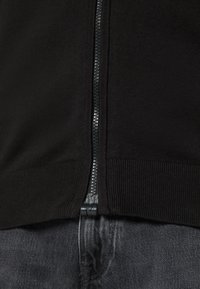Sweat à zip noir avec ourlet et poignets côtelés, en tissu tricoté texturé. La fermeture éclair est noire avec un tirant en argent.