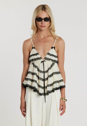 Donna bionda che indossa occhiali da sole neri e un top crema senza maniche con volant in pizzo nero e piccoli fiocchi neri, abbinato a pantaloni crema.