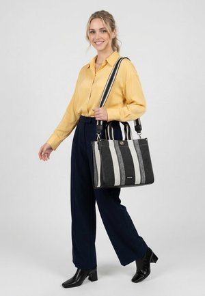 Femme portant une chemise jaune et un pantalon bleu marine, tenant un sac fourre-tout à rayures noires et blanches, souriante, debout devant un fond uni.