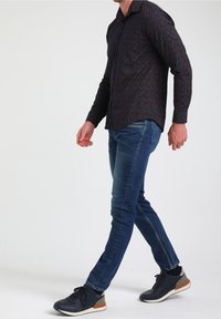 Camicia nera con bottoni, decorata con un motivo geometrico, abbinata a jeans blu aderenti e sneakers nere con un accento beige.