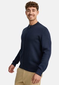 Suéter azul marino con cremallera y cuello redondo, puños y bajo acanalados. Textura suave y diseño ajustado, usado sobre pantalones beige.