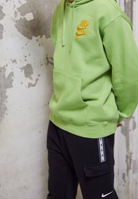 Grön bomullssweatshirt med gul Nike-logotyp, känguruficka och dragsko. I kombination med svarta byxor med sidogylf och logotypdetalj.