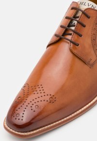 Chaussure en cuir poli marron avec un bout pointu, un détail perforé complexe et des lacets marron, présentant une couture blanche contrastante le long de la semelle.