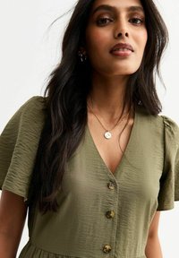 Vestido verde oliva con escote en V, mangas cortas y tela texturizada, que presenta tres botones decorativos en el frente.