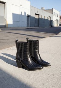 Paar schwarze knöchelhohe Lederstiefel mit Krokodilstruktur und Blockabsätzen, die bei Tageslicht auf einem städtischen Gehweg stehen.