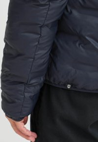 Marinblå pufferjakke med en glat, skinnende overflade, der har en quiltet tekstur og et logo-tag i kanten. Ærmet er taget fra bagfra.