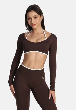 Młoda kobieta ubrana w brązowy crop top z długim rękawem i dopasowane legginsy z białym wykończeniem, stojąca na jednolitym tle.