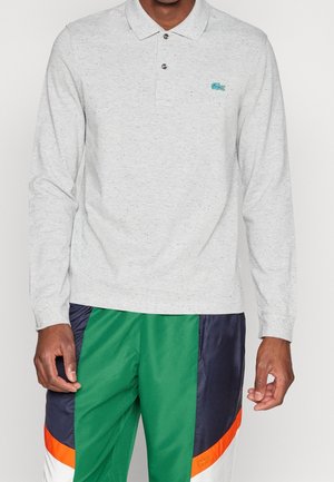 Grijze poloshirt met lange mouwen en een gespikkelde textuur, drie knopen en een groen geborduurd logo op de borst. Gecombineerd met kleurrijke sportieve broek.