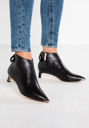 Botines de cuero negro con punta afilada, cremallera de color oro rosa y detalles en el tacón, combinados con jeans ajustados azules.
