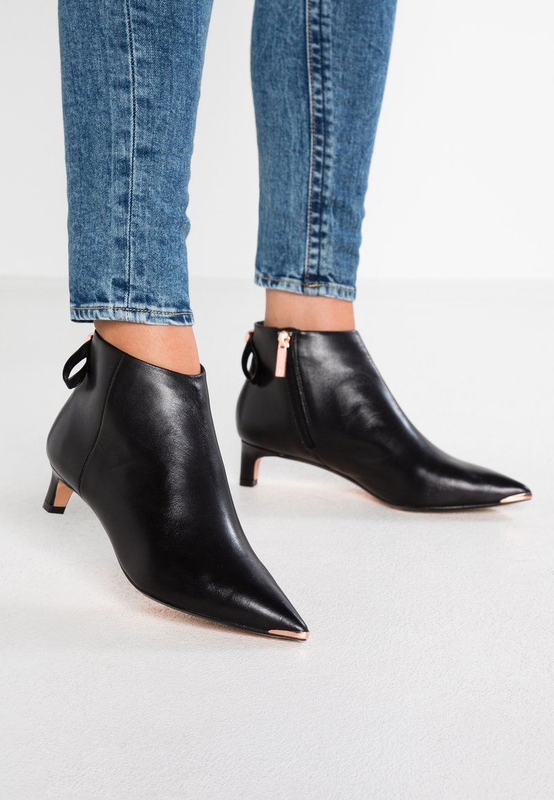Bottes chevilles pointues en cuir noir avec fermeture éclair et talons en or rose, portées avec un jean skinny bleu.