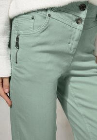 Pantalones verde claro con un bolsillo con cremallera, cierre de botón y tejido texturizado. Las características incluyen costuras en contraste y detalles metálicos.