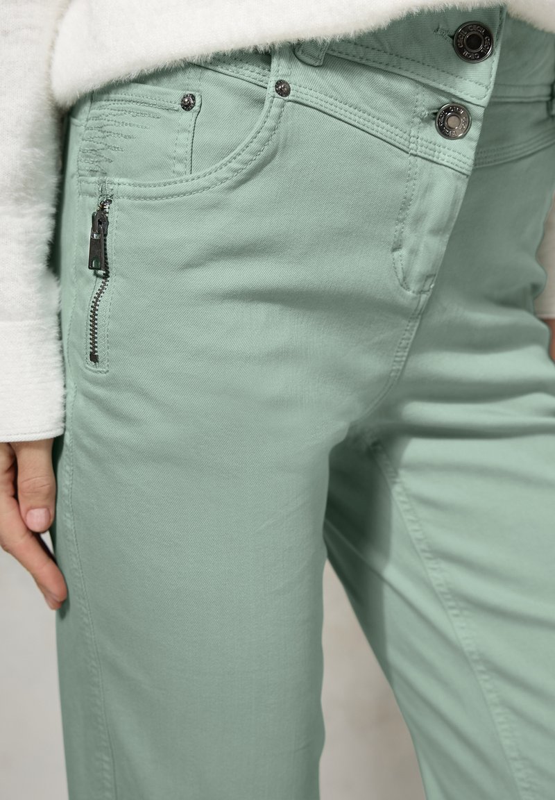 Pantalones verde claro con un bolsillo con cremallera, cierre de botón y tejido texturizado. Las características incluyen costuras en contraste y detalles metálicos.