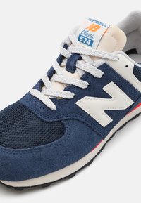 Baskets navy avec tige en daim et mesh, logo en cuir blanc, lacets texturés et languette crème. Accent rouge sur la semelle. Design classique.
