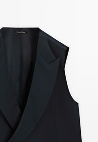 Schwarzer maßgeschneiderter ärmelloser Blazer mit breitem Revers und einem "Personal Tailoring"-Label am Kragen, vor einem weißen Hintergrund abgebildet.