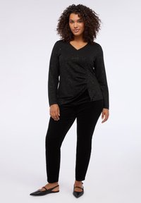 Black long-sleeve top with a V-neck and textured metallic sparkles; features a side ruching detail. Paired with black fitted pants and flat shoes.