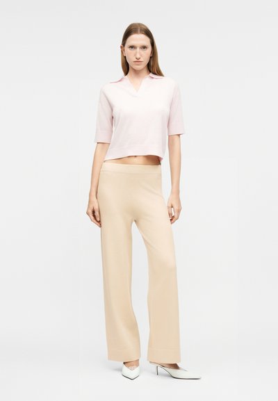 Γυναίκα που στέκεται φορώντας ένα ανοιχτό ροζ μπλουζάκι με κοντά μανίκια και γιακά, μπεζ παντελόνι με φαρδιά κουμπιά και λευκά μυτερά τακούνια σε λευκό φόντο.