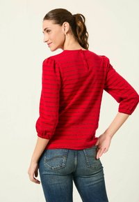 Rotes Oberteil mit horizontalen schwarzen Streifen, featuring Puffärmel und einer Knopfleiste am Rücken. Kombiniert mit blauen Jeans.