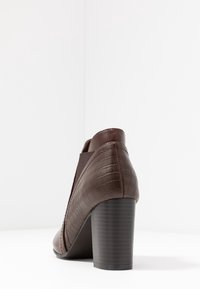 Botín de tobillo marrón con tacón, hecho de cuero texturizado con un patrón de cocodrilo. Presenta un tacón negro sólido y paneles laterales elásticos.