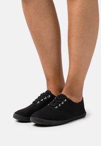 Czarne sneakersy na płaskiej podeszwie, z białymi oczkami i czarnymi sznurówkami. Teksturowana gumowa podeszwa zapewniająca przyczepność i trwałość.