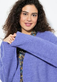 Lila stickad cardigan med ribbad manschett; mjuk textur; lagd över en blommönstrad topp i blå och gyllene nyanser.